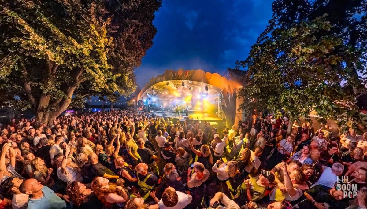 Enthousiast festivalpubliek voor een openlucht podium bij schemering, bomen omlijsten het verlichte podium met The Dutch Queen
