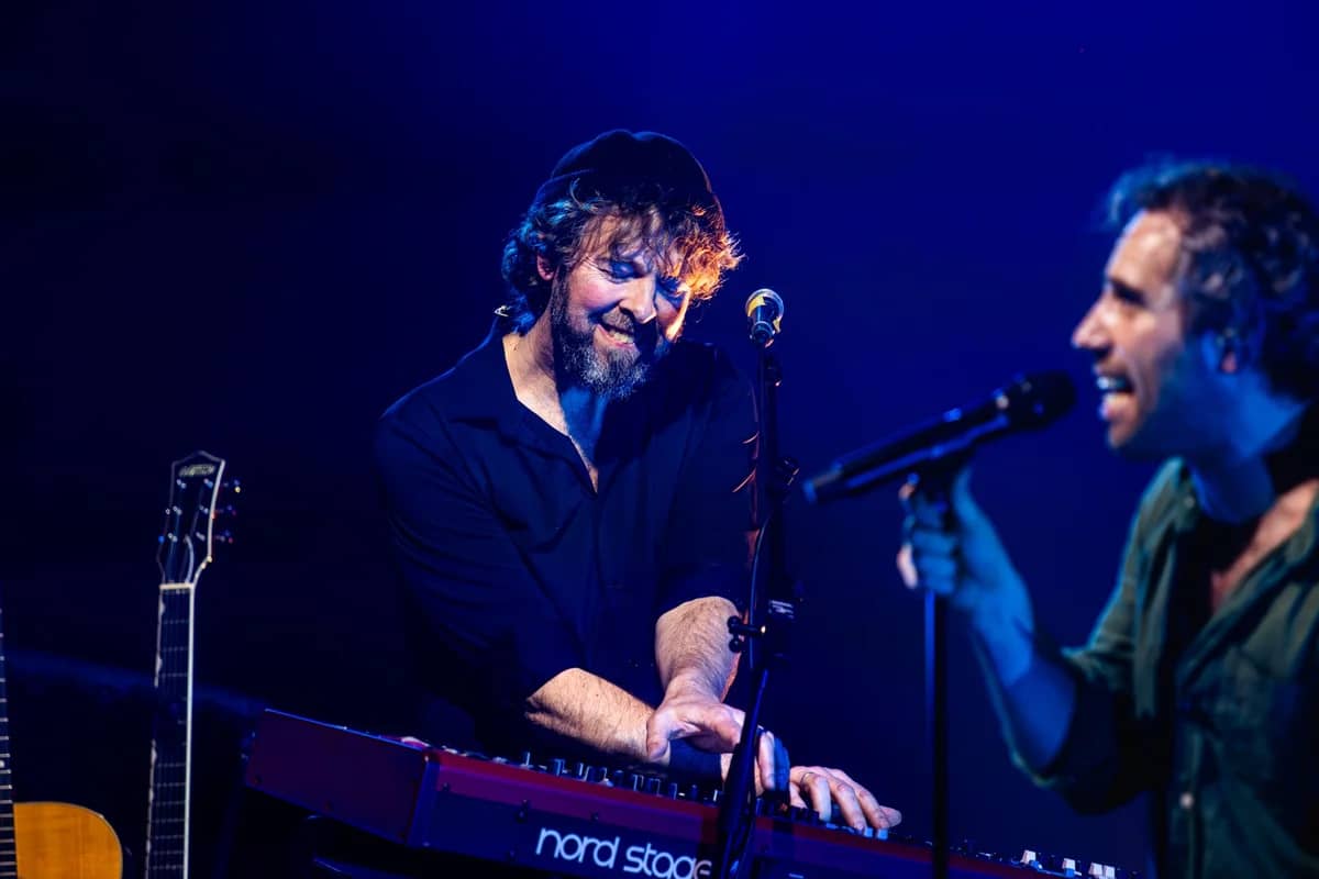 Toetsenist speelt lachend op een Nord Stage keyboard naast de zanger, intiem moment in blauw podiumlicht