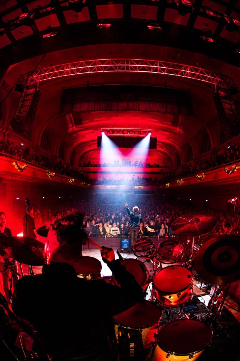 Uitzicht vanaf het drumstel over een uitverkocht theater baadend in rood licht, zanger interacteert met het publiek